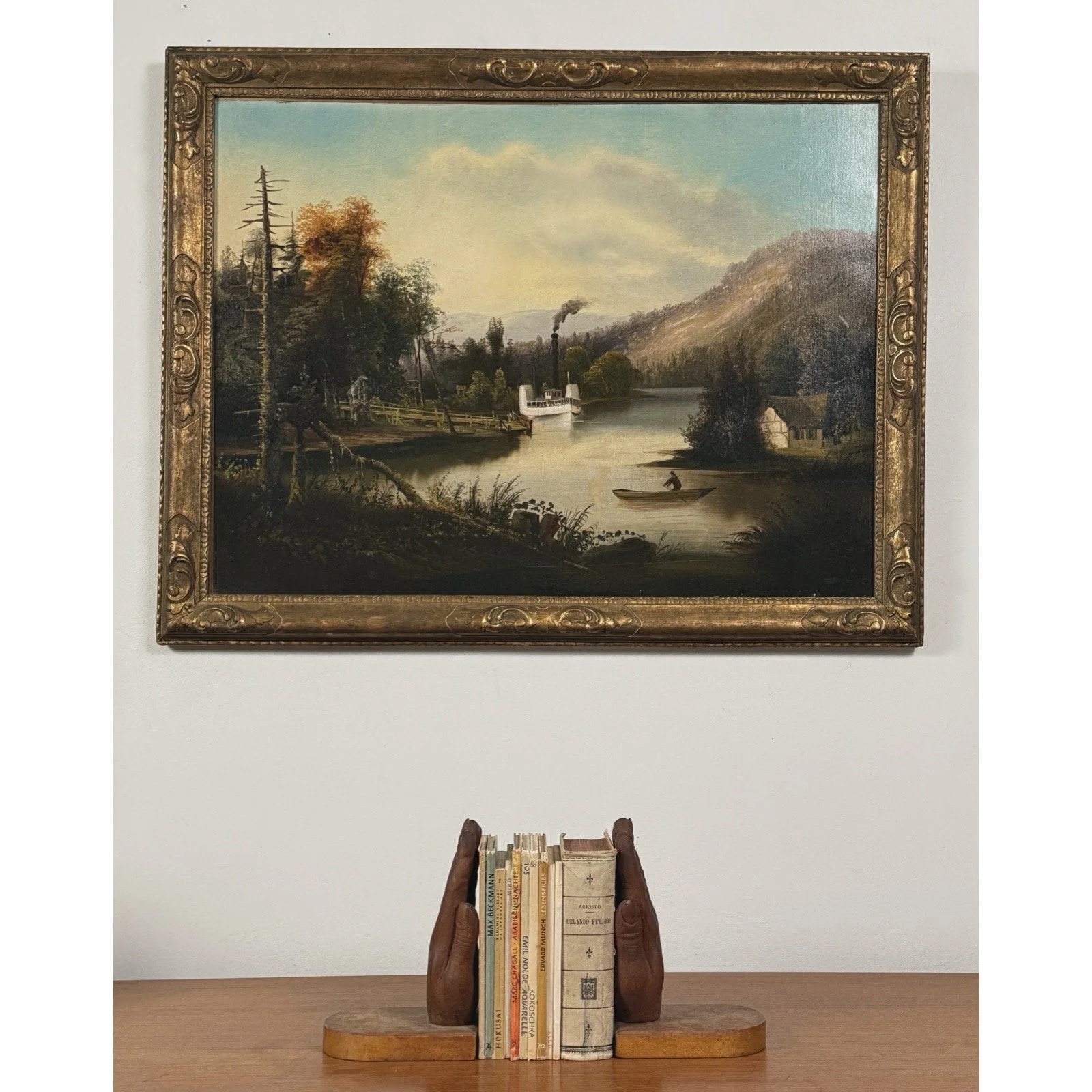 Full frontal view of an antique American folk art oil painting depicting a river landscape with steamboat and rowboat with wood bookends.