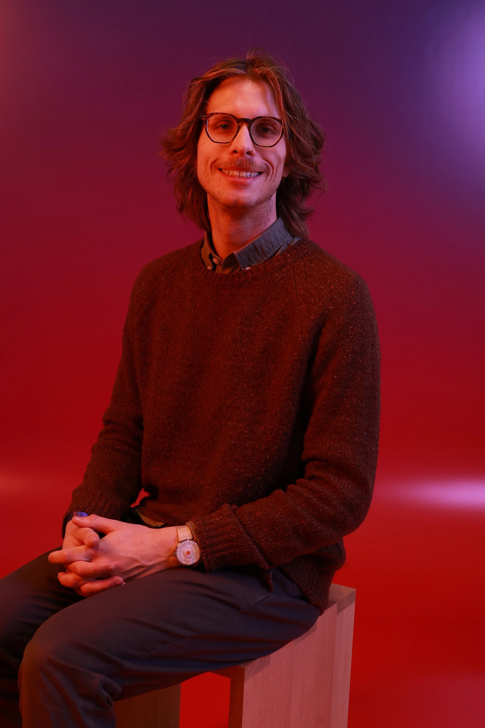 A man with shoulder-length brown hair, glasses, and a short mustache, smiling, sitting on a wooden stool against a colorful background with pink, purple, and red hues, wearing a dark red sweater and dark pants.