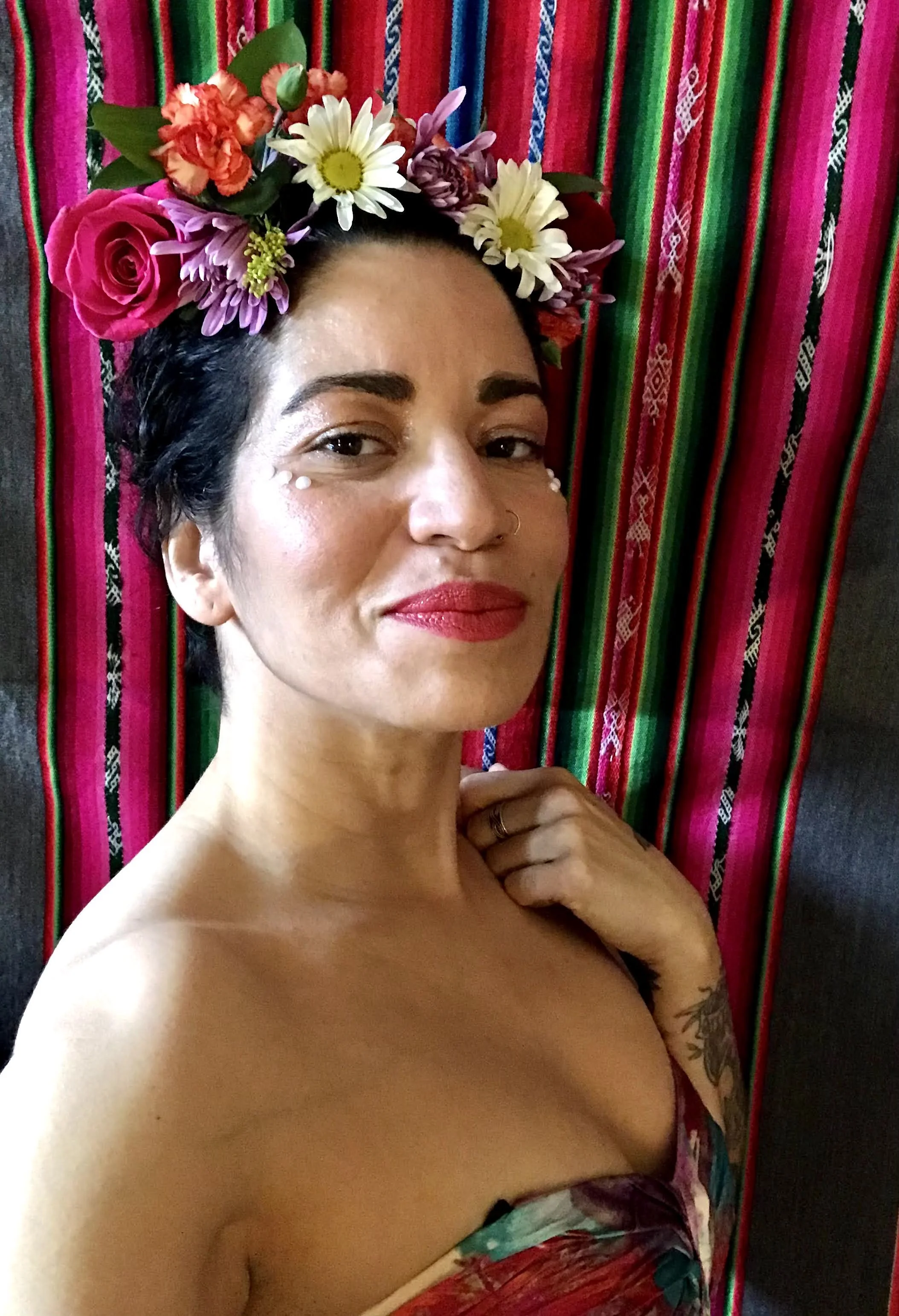 A woman with dark hair, red lipstick, and a flower crown of roses, daisies, and other flowers, standing against a colorful striped textile background. She has makeup with small decorative white dots below her eyes, a nose ring, and a tattoo on her left arm.