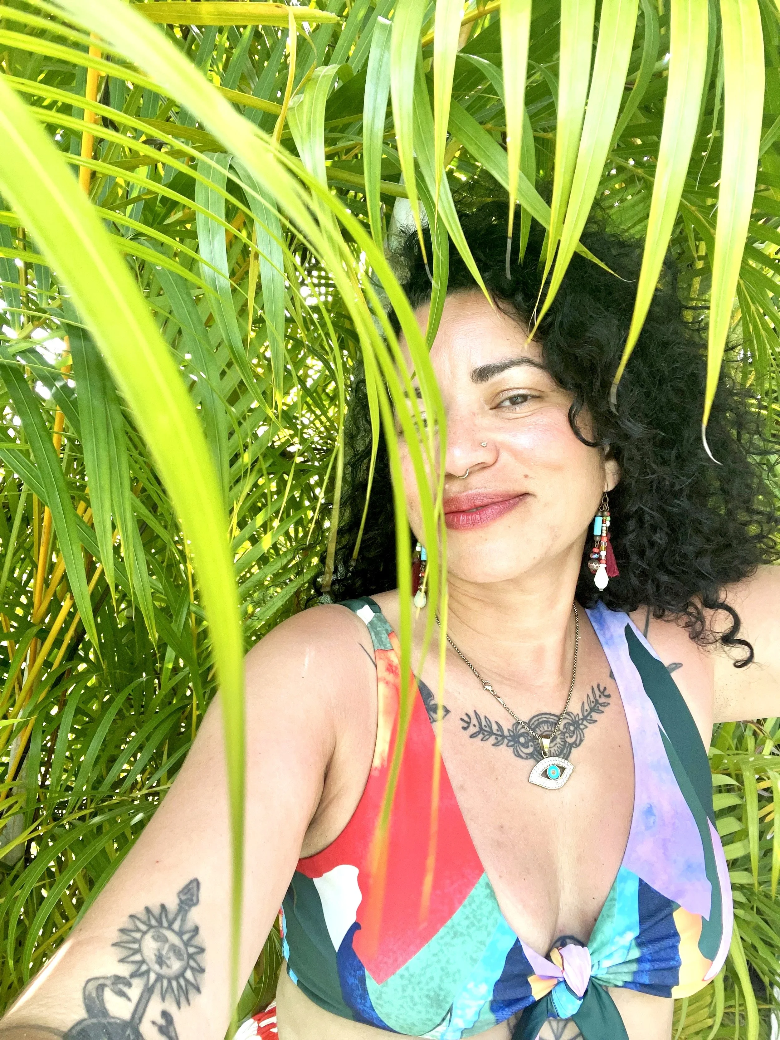 Artist with curly black hair smiling, wearing colorful tie-dye swimsuit, surrounded by green palm leaves.