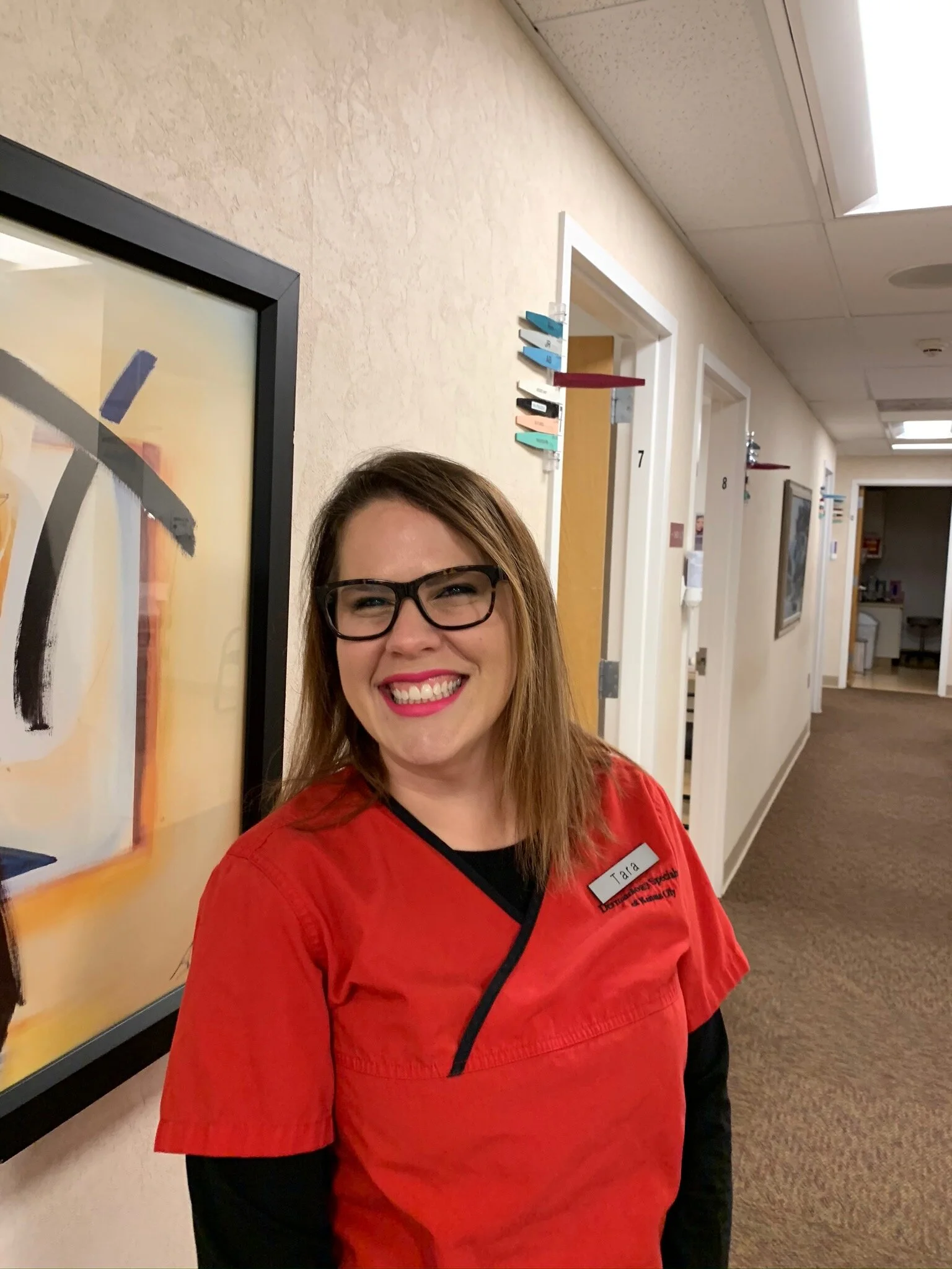 A smiling woman with glasses and red medical scrubs standing in a corridor of a healthcare facility.