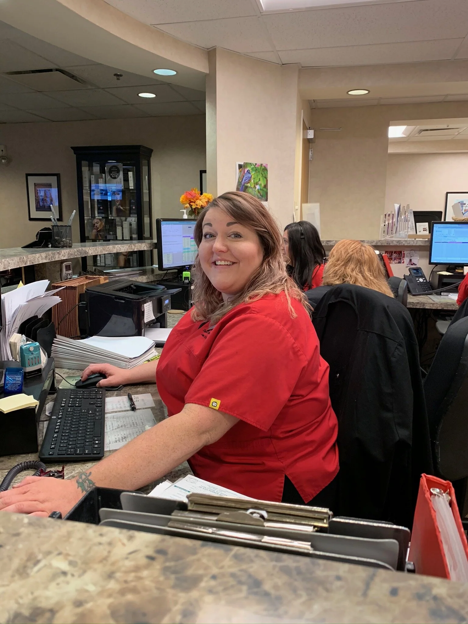 A smiling woman in a red uniform sitting at a desk with a computer in an office environment, with other employees working behind her.
