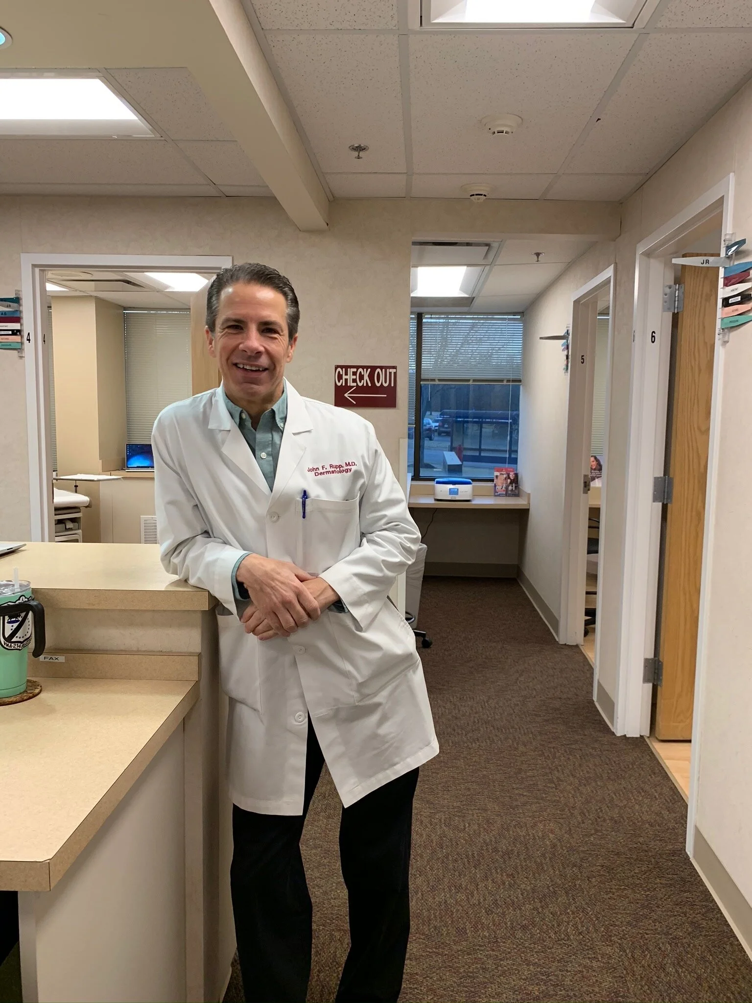 A male doctor in a white coat standing at a reception desk in a clinic or hospital office, smiling at the camera.