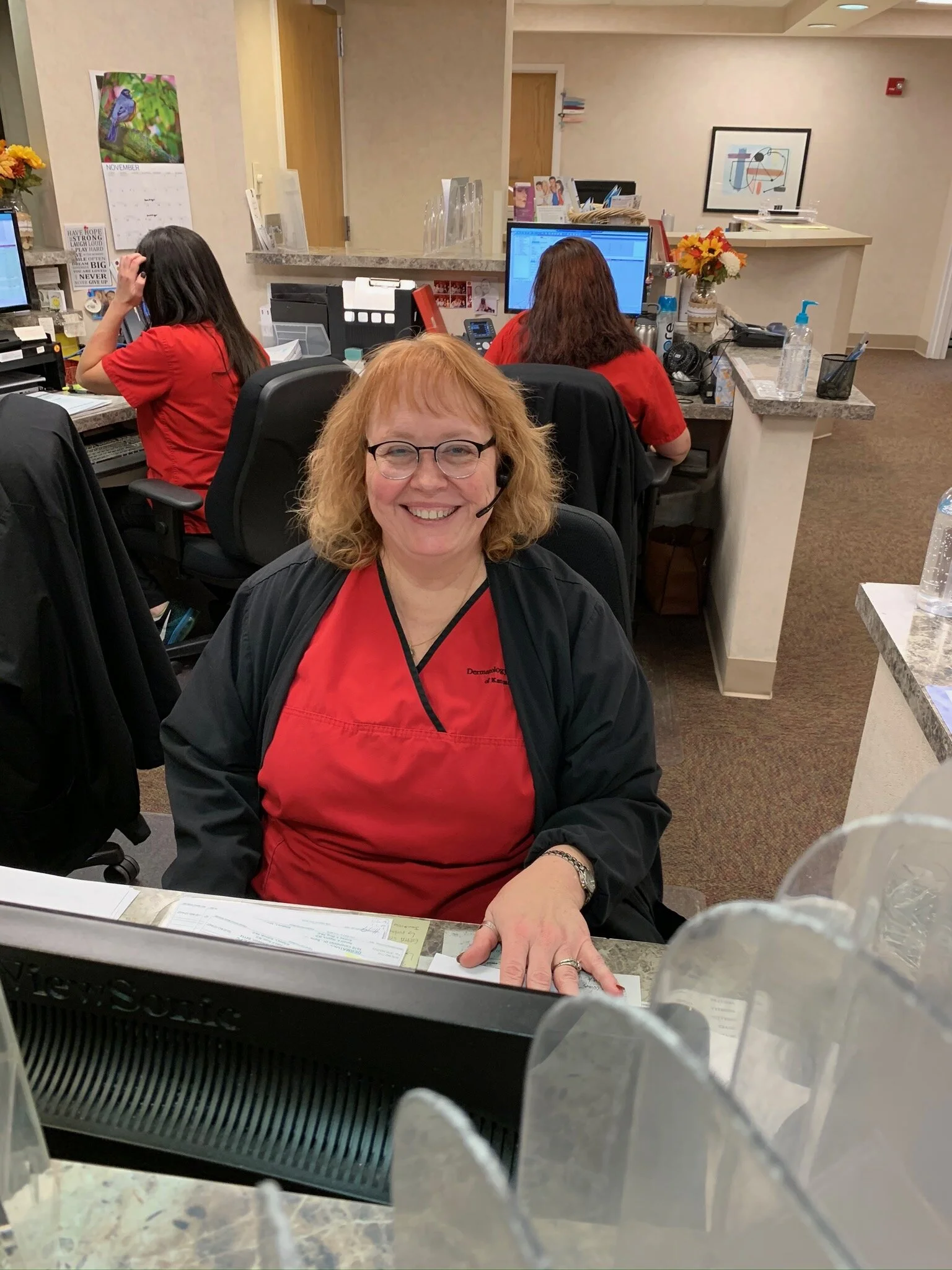 A woman with red hair, glasses, and wearing a red uniform with a headset, smiling at a desk in an office setting. Behind her are two other women working at computers, also in red uniforms.
