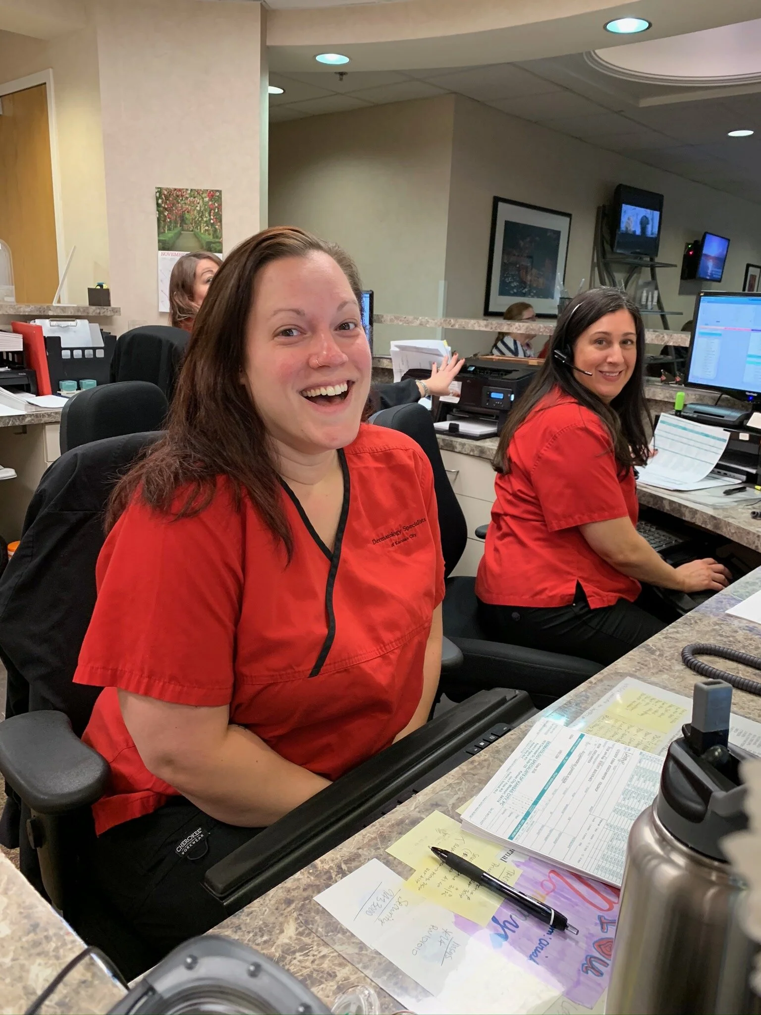 Two women in red uniforms working at a reception or administrative desk in a medical facility. One is smiling at the camera, and others are working in the background.