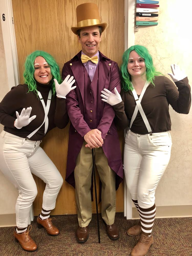 Three people dressed in costumes, two women with green hair and white striped socks, and a man in a purple coat and tall gold hat, standing in front of a wooden door.