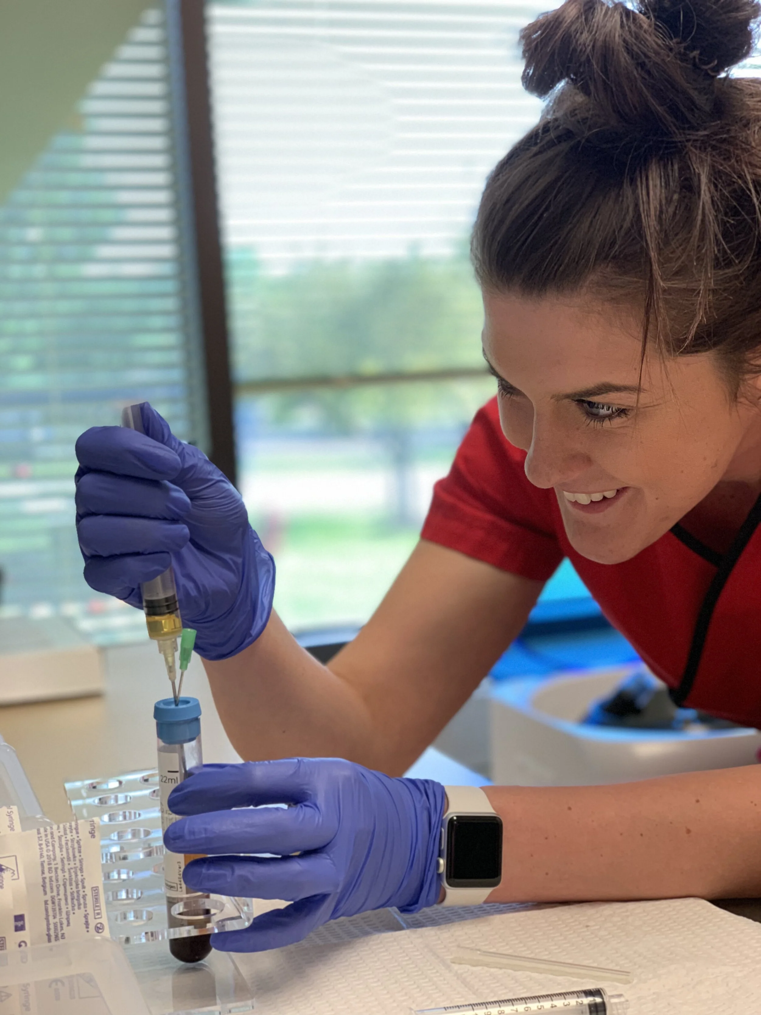 A woman with dark hair in a bun wearing a red shirt and blue gloves drawing blood from a person at a medical testing site.