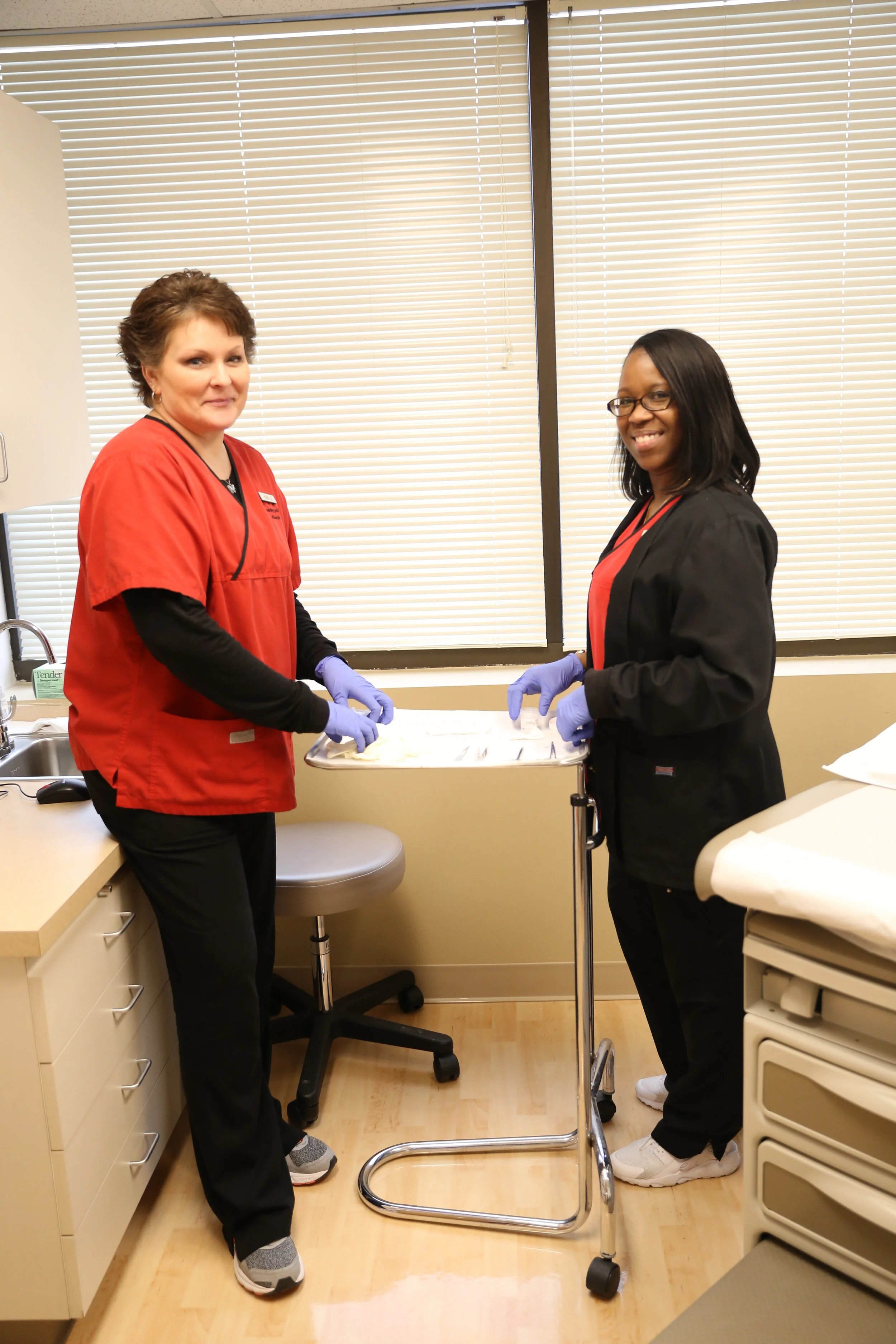 Two healthcare workers in scrubs and gloves standing in a hospital room, smiling, next to a medical cart and a bed.