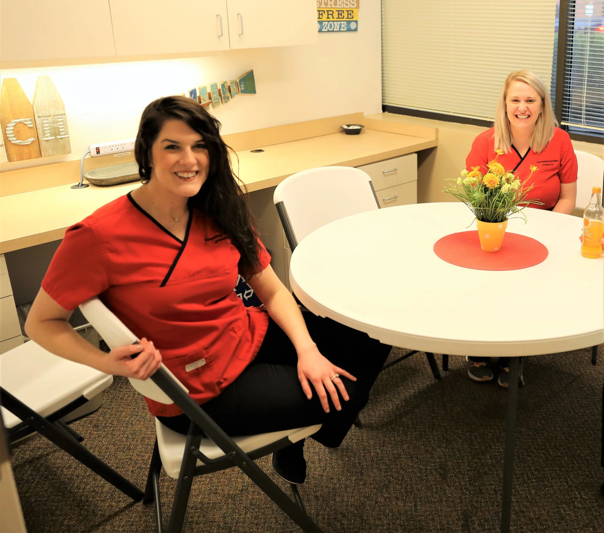 Two women in red scrubs smiling in a room with a round table, a flower pot with yellow flowers, and a soda bottle, with window blinds and decor on the wall.