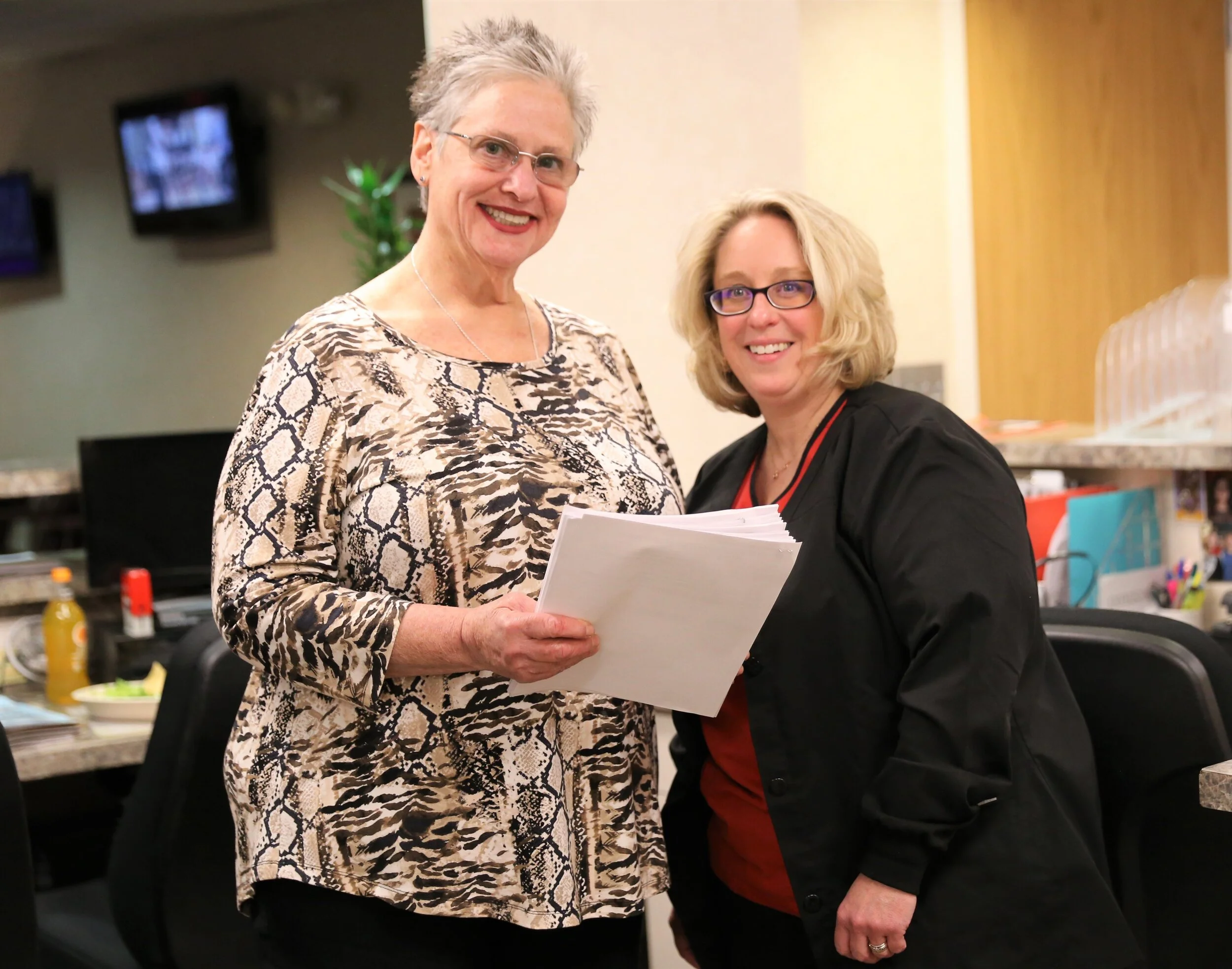 Two women smiling and holding papers in an office setting, with desks, chairs, and a computer in the background.