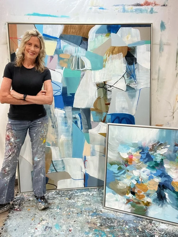 A woman standing with crossed arms, smiling, in an art studio next to two abstract paintings with blue, white, and earth-tone colors, on a splattered art floor.