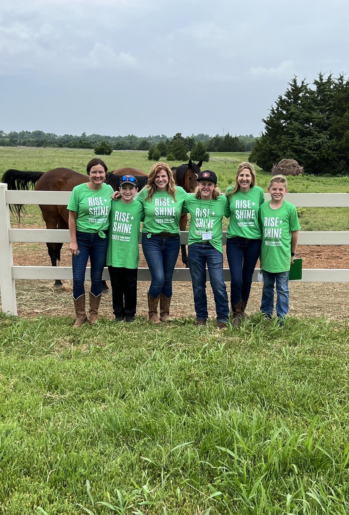 Group of people wearing green "Rise + Shine Camp" shirts in front of fence, two large brown horses stand behind them.