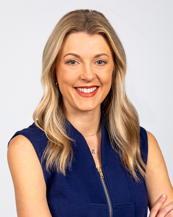 A woman with blonde hair wearing a navy blue sleeveless dress, holding a teal speech bubble sign with a white outline of a smiling face in a speech bubble. She is smiling at the camera with a white background and a blue rectangular design behind her.