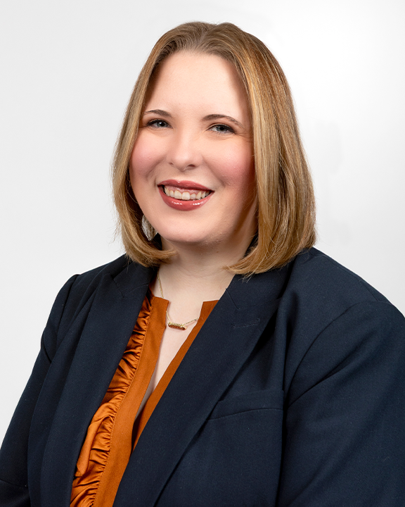 A woman with shoulder-length red hair, wearing a plaid dress with a bow tie, holding a teal speech bubble sign with a white smiley face icon, smiling at the camera.