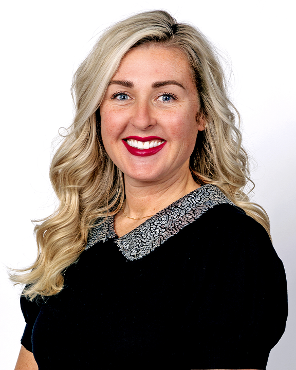 A woman with blonde hair smiling, holding a green speech bubble sign with a white child's handprint. She has a name badge and a teal badge on her sweater.