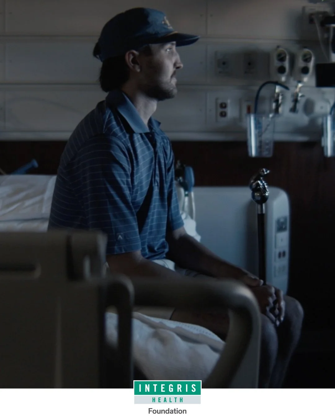 A man sits on a hospital bed in a dimly lit hospital room, wearing a blue cap and hospital gown, looking to the right with a contemplative expression.