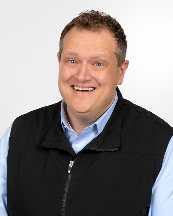 A man smiling and holding a sign with a speech bubble shape featuring a stylized baby icon, wearing a dark blue jacket and a watch.