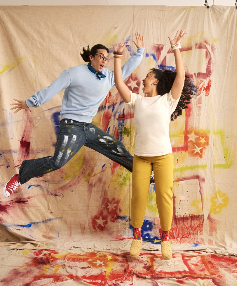 Two women are smiling and celebrating in front of a colorful, abstract backdrop, with one jumping mid-air and the other raising her arms.