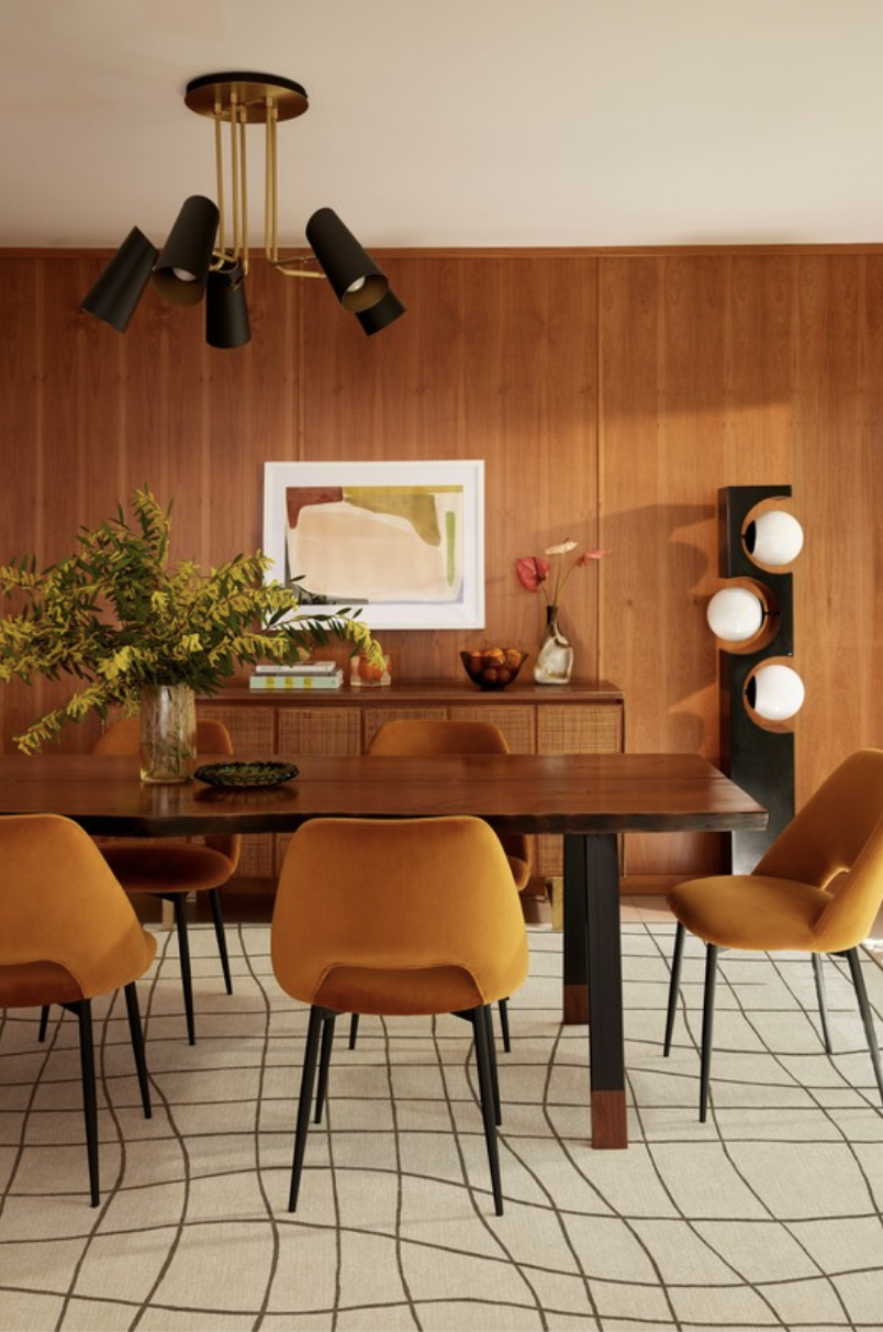 A dining room with a wooden table surrounded by mustard-colored chairs, a sideboard with decorative items, a modern black and white light fixture on the ceiling, and a wooden-paneled wall with abstract art and a unique lighting sculpture.