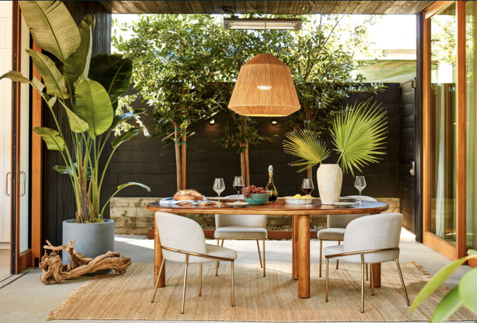 A dining area with a wooden table set for a meal, surrounded by white upholstered chairs, next to large potted plants and greenery outside large open doors.