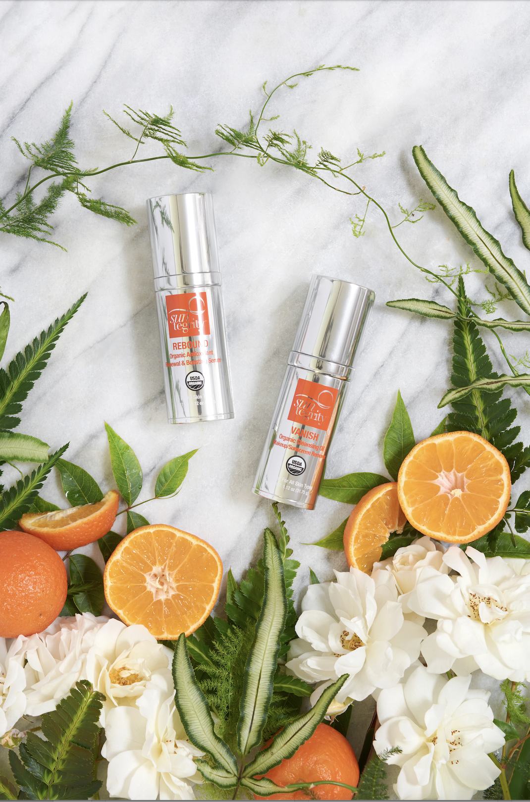 Two skincare bottles labeled 'Rebound' and 'Vanish' surrounded by orange slices, white flowers, and green leaves on a white marble background.