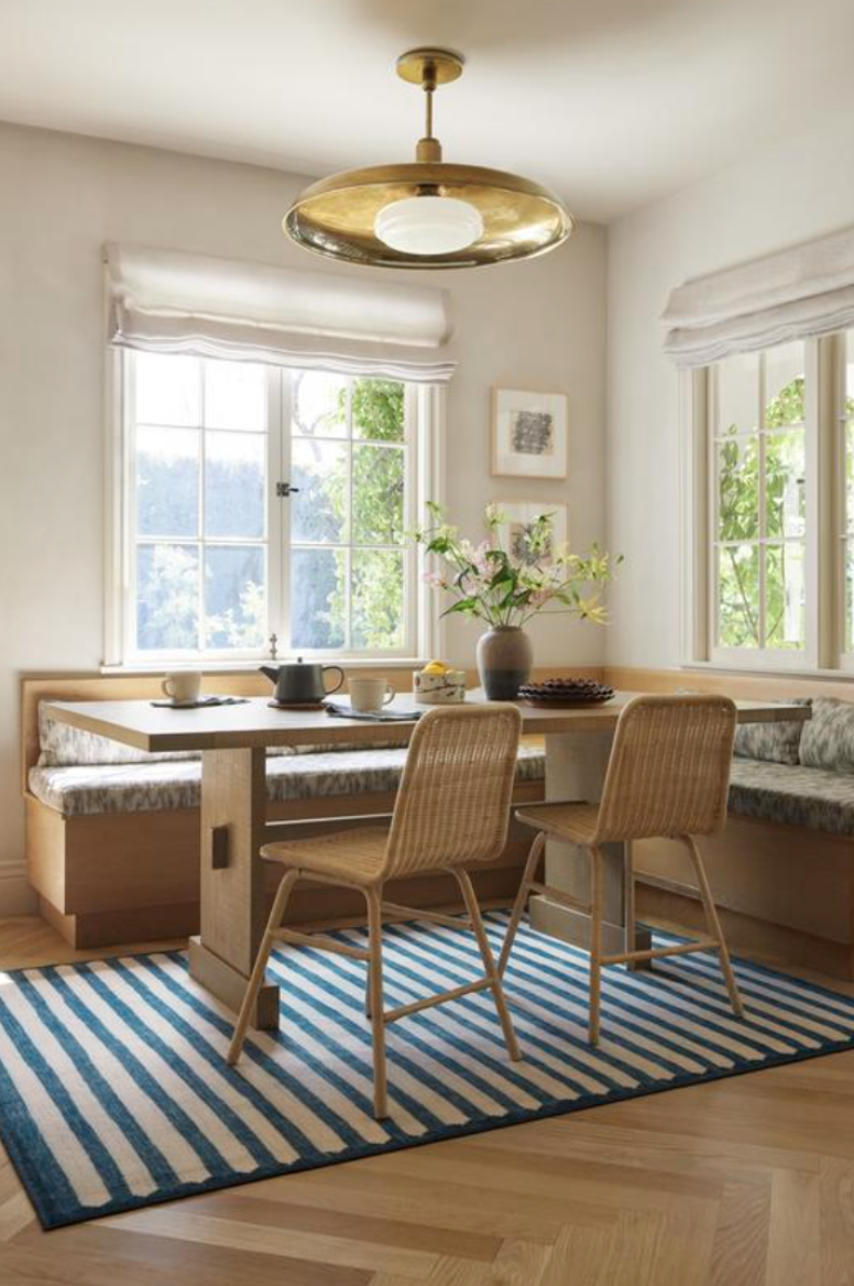 A cozy dining nook in a well-lit room with two large windows, a wooden table, two wicker chairs, a built-in bench with cushion, a blue and white striped rug, a vase with flowers on the table, and a ceiling light fixture.