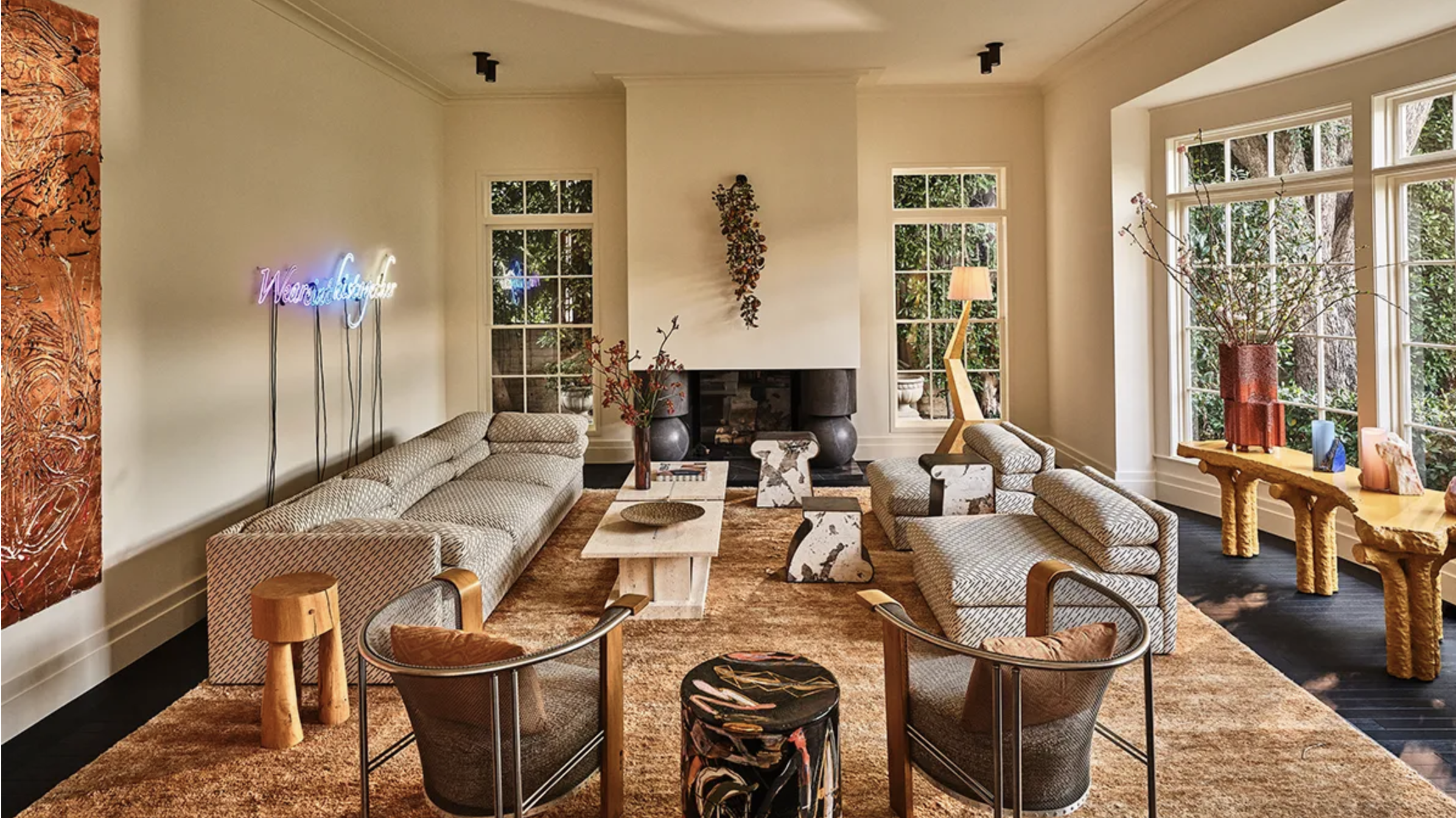 A cozy living room with large windows, beige sofas, a coffee table, artwork, plants, and decorative lighting, featuring a warm and inviting atmosphere.