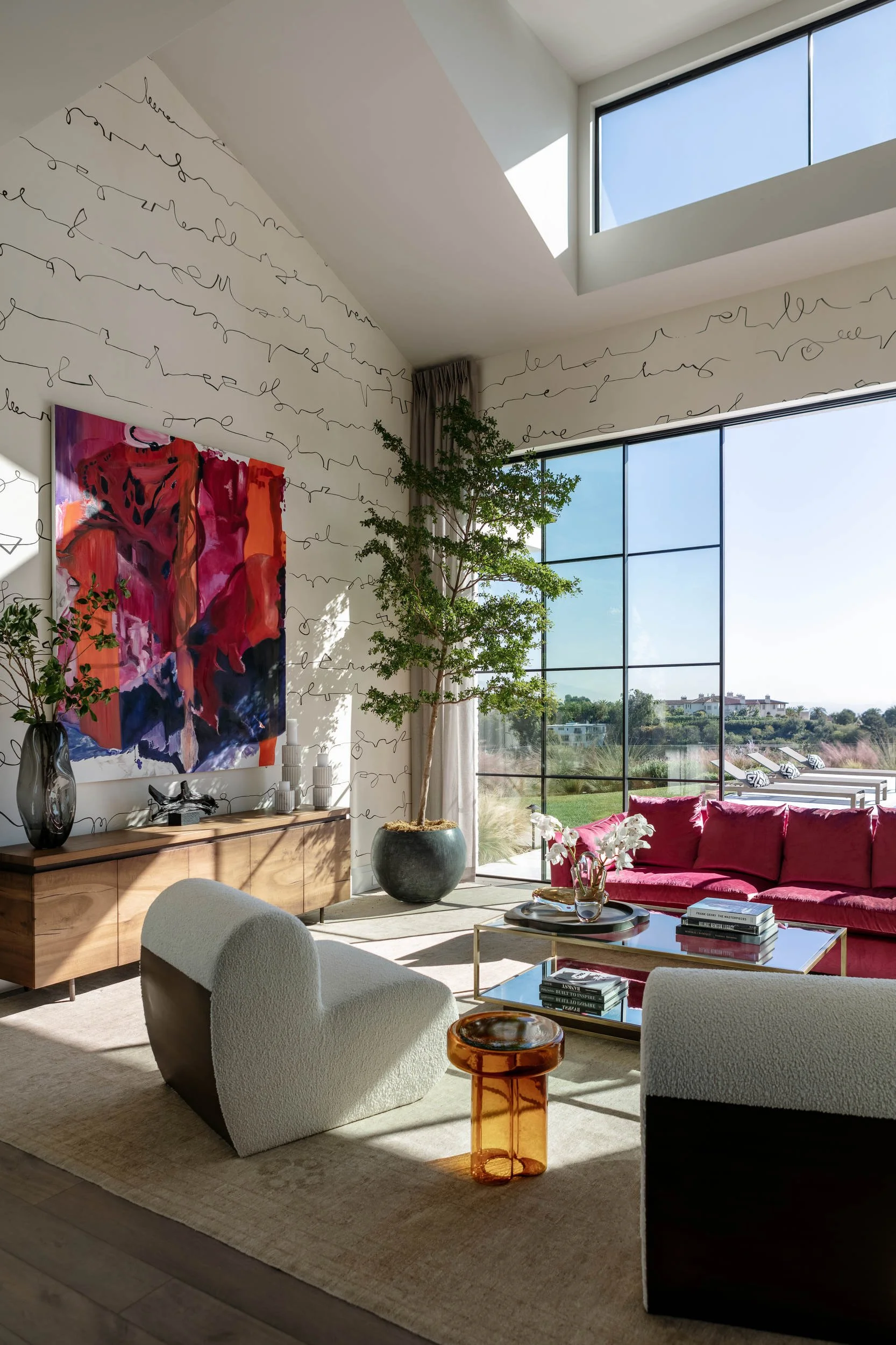 Modern living room with high ceilings, large window, red sofa, white and black chairs, abstract art, small side table, potted tree, and books.