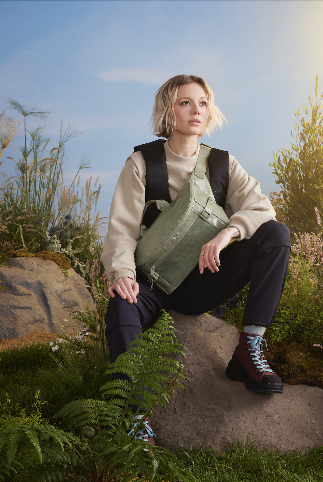 A woman sitting outdoors on a rock surrounded by greenery and flowers, wearing a beige sweatshirt, black pants, red hiking boots, and carrying a green bag with a black backpack strap.