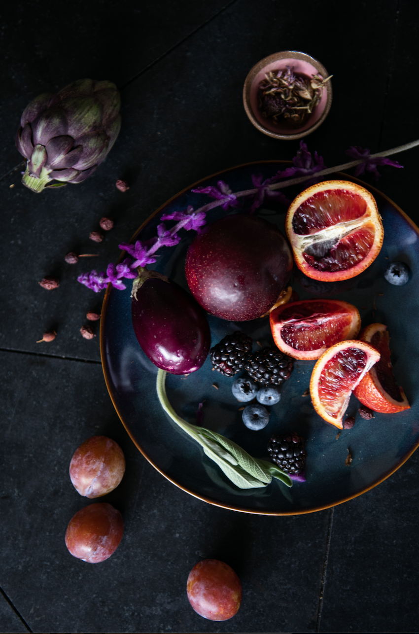 A dark plate containing blood oranges, blackberries, blueberries, and an eggplant, with additional fruits and a purple artichoke on a dark surface.
