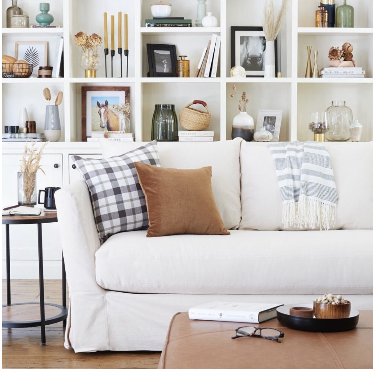 Living room with white sofa, checkered and solid brown pillows, bookshelf with decor and books, side table with black mug, glasses, and a book, and a coffee table with tray, glasses, and a book.