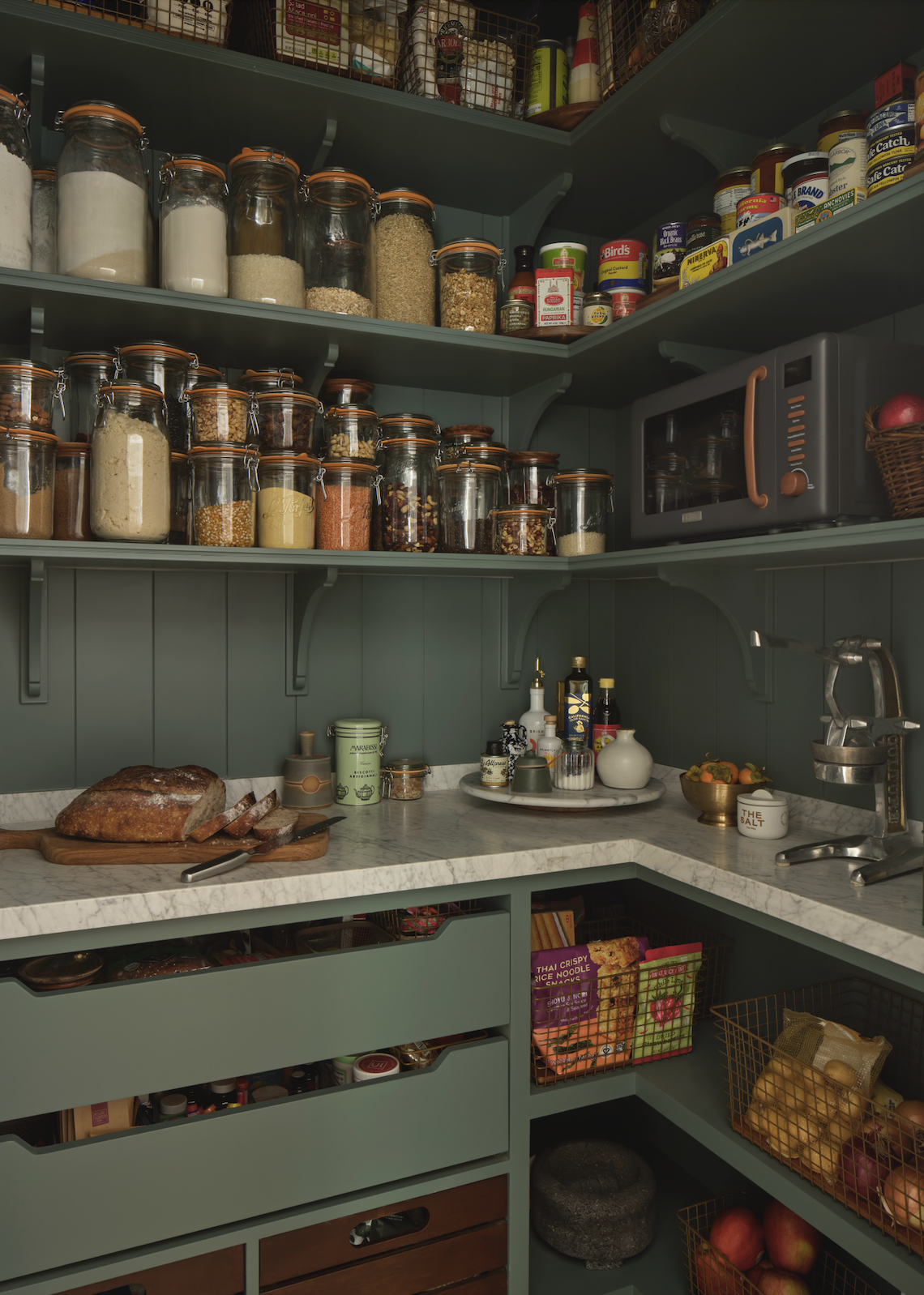 A well-organized kitchen pantry with green cabinets, glass jars filled with dry ingredients, canned foods, a microwave, a cutting board with a loaf of bread, and various kitchen essentials on the marble countertop.