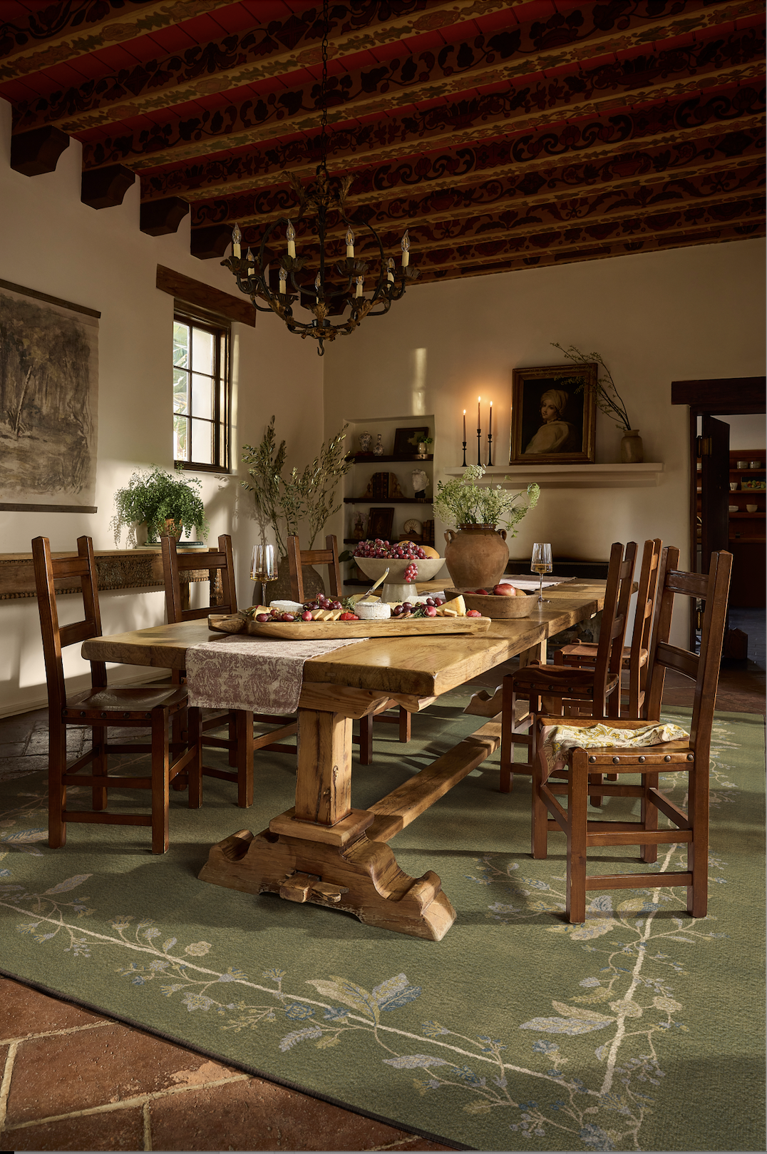 A rustic dining room with a wooden table set on a green patterned rug, surrounded by wooden chairs. The table has a large vase with greenery, cutlery, and bowls of fruit. The ceiling features exposed wooden beams with decorative painted patterns, and a chandelier hangs overhead. Sunlight streams through a window, illuminating the cozy and traditional decor, including a portrait on the wall and a small bookshelf.
