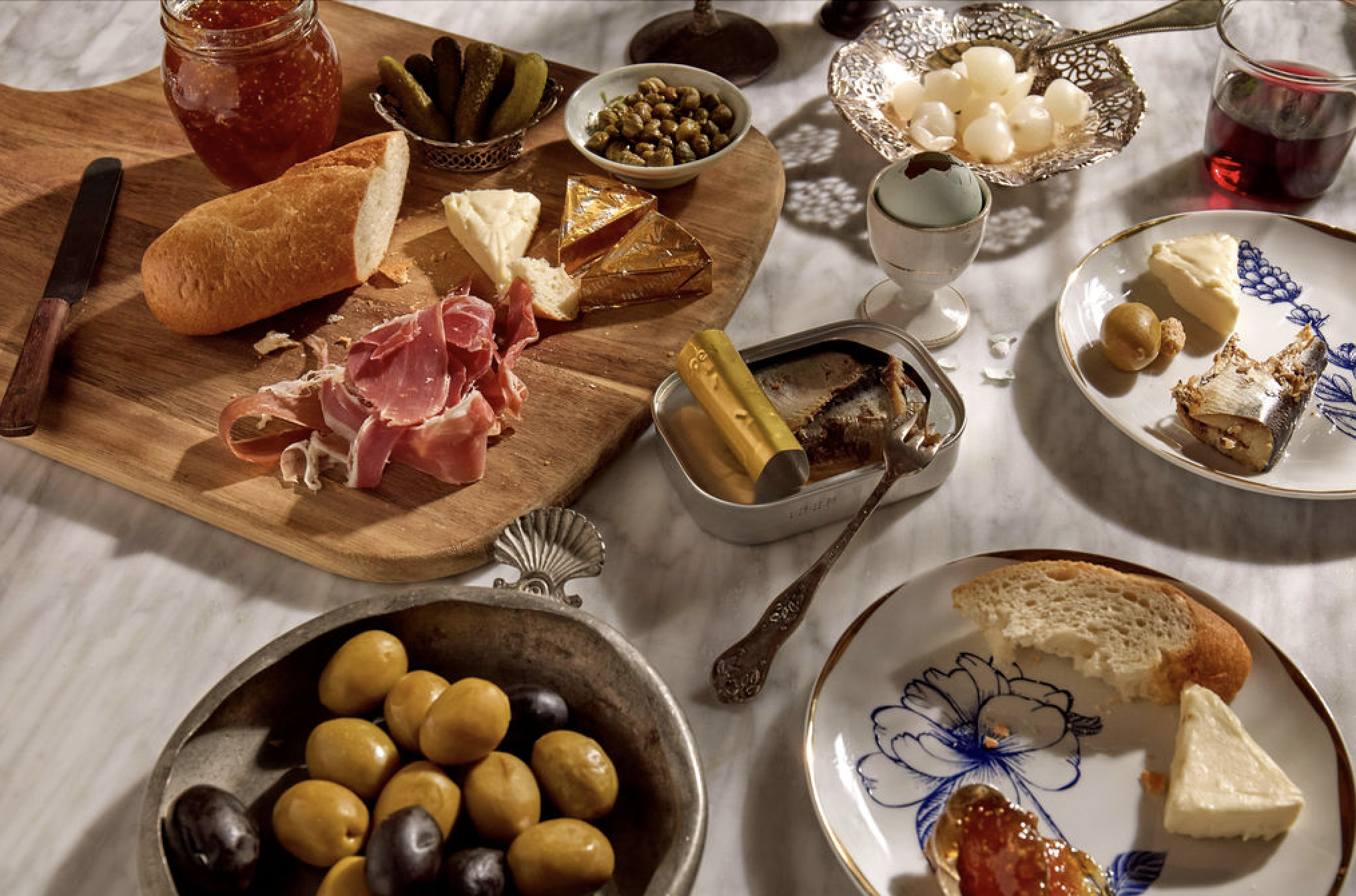 A cheese and charcuterie board with bread, pickles, olives, and various cheeses, surrounded by small bowls of jams, fruit, and wine on a table.