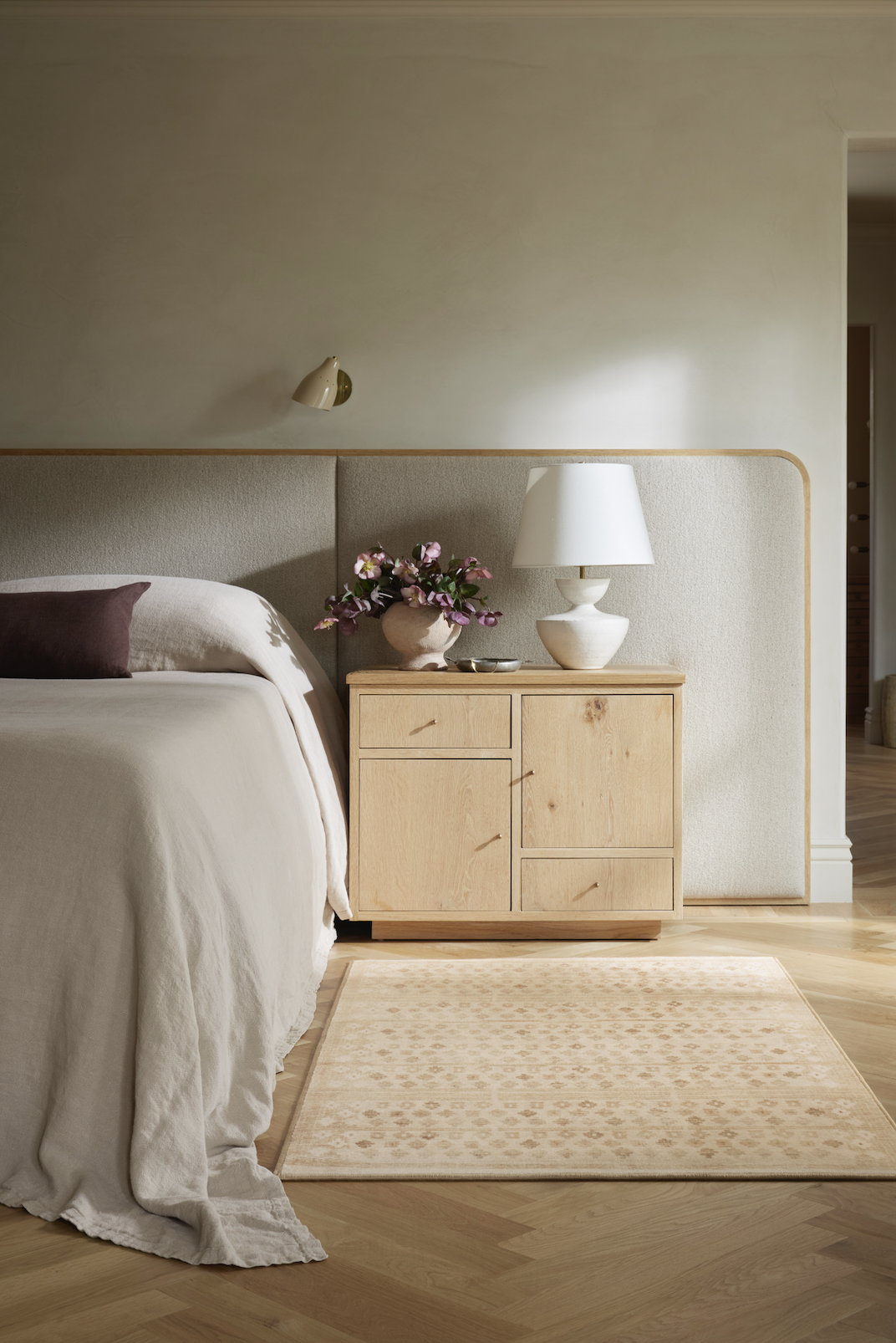 A bedroom with a beige upholstered headboard, a nightstand with a white lamp and pink flower arrangement, and a beige rug on light wood flooring.