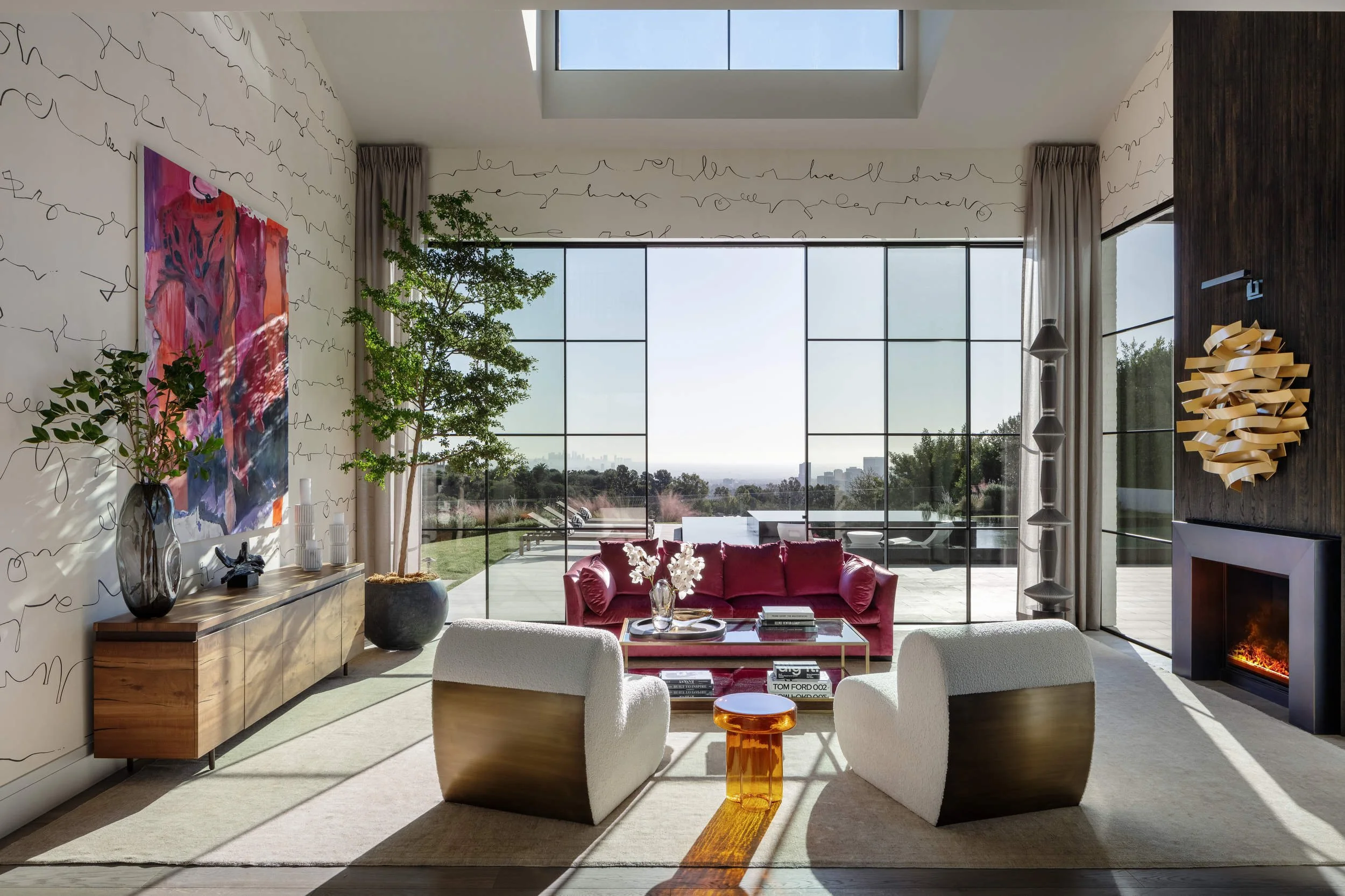Modern living room with large floor-to-ceiling windows, a red sofa, white armchairs, abstract wall art, decorative objects, a potted plant, and a view of the outdoors.