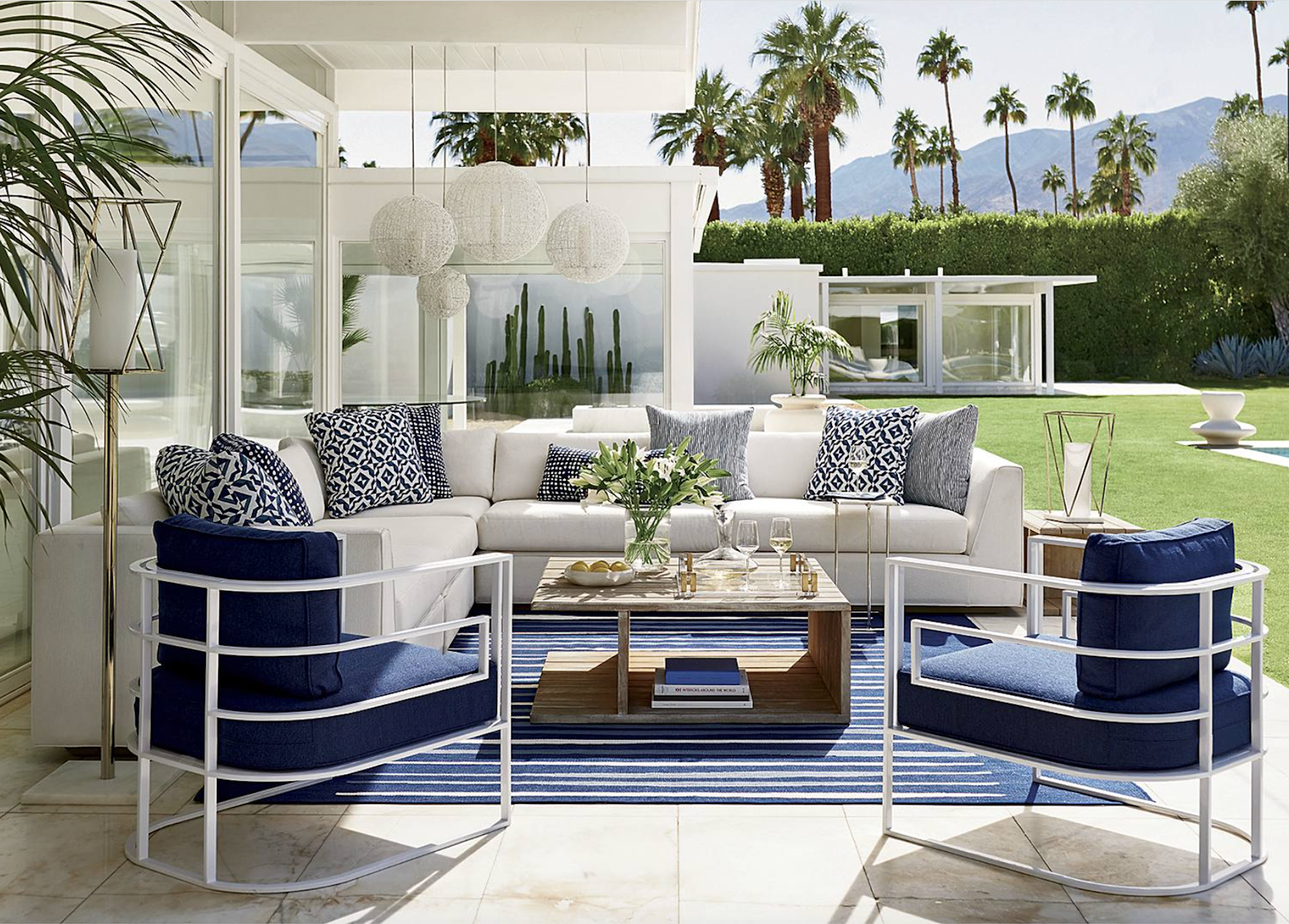 Outdoor patio furniture with a white sectional sofa, navy and patterned pillows, and two navy chairs with white frames, on a blue and white striped rug, with a wooden coffee table holding a vase of flowers and glasses, set against a backdrop of palm trees and mountains.