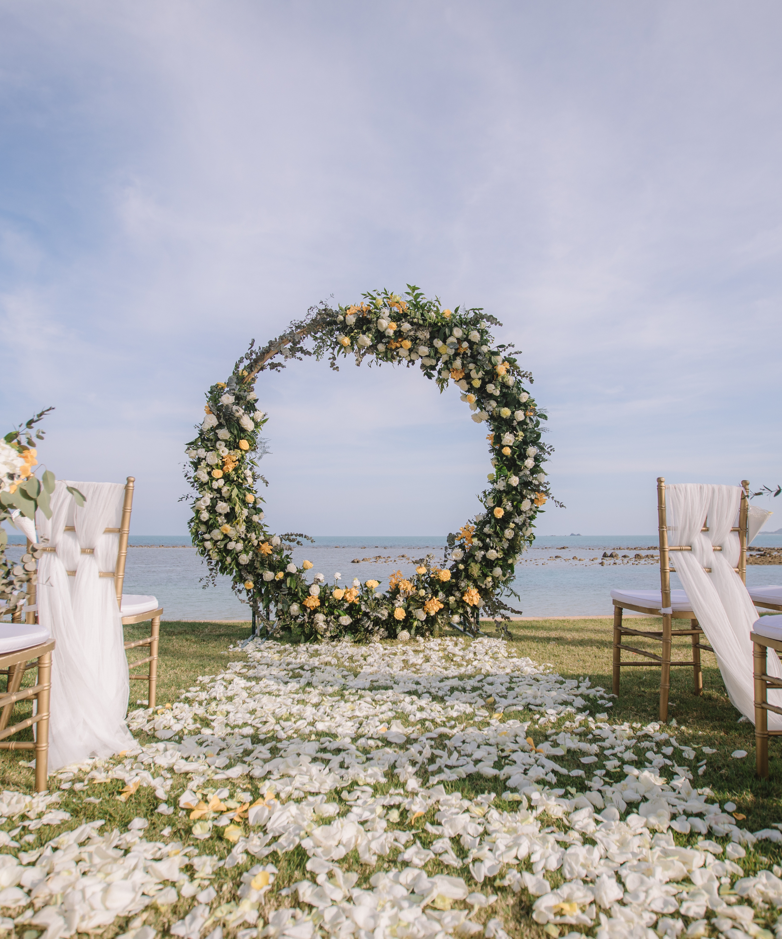 A beachside wedding ceremony setup with a floral arch and white chairs, covered in white flower petals, overlooking the ocean and sky.