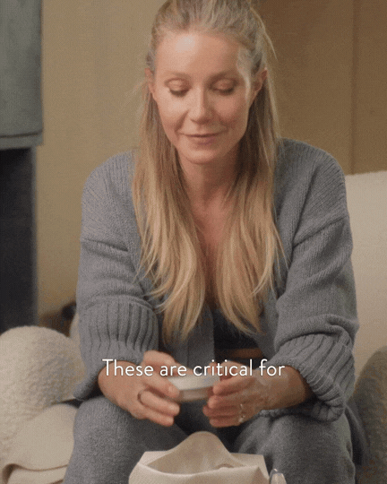 A woman with long blonde hair wearing a gray sweater, sitting indoors and looking down at her hands.