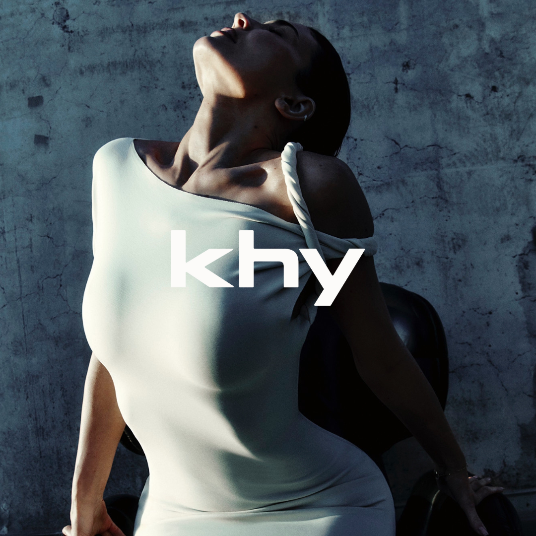 A woman in a white dress posing against a textured gray wall, with her head tilted back and eyes closed, partially illuminated by natural light.