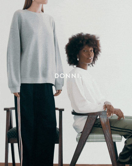 Two women in casual clothing, one standing and one sitting, in a minimalist room with chairs. The standing woman wears a gray sweatshirt and black track pants, while the seated woman wears a white sweater and olive-green pants. The word 'DONNI.' is visible in the middle of the image.