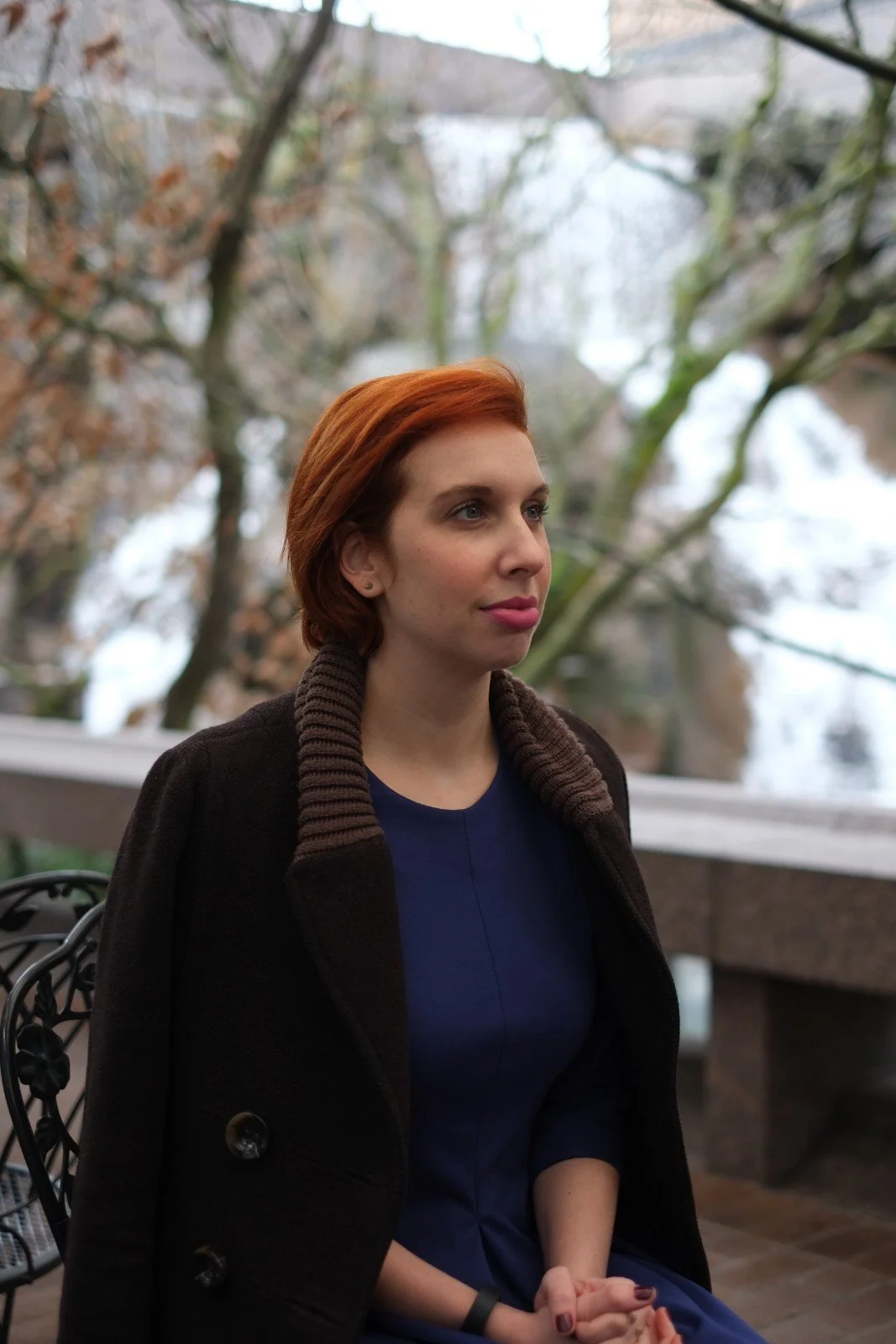 A woman with short red hair sits with her hands clasped in tree-filled urban setting.