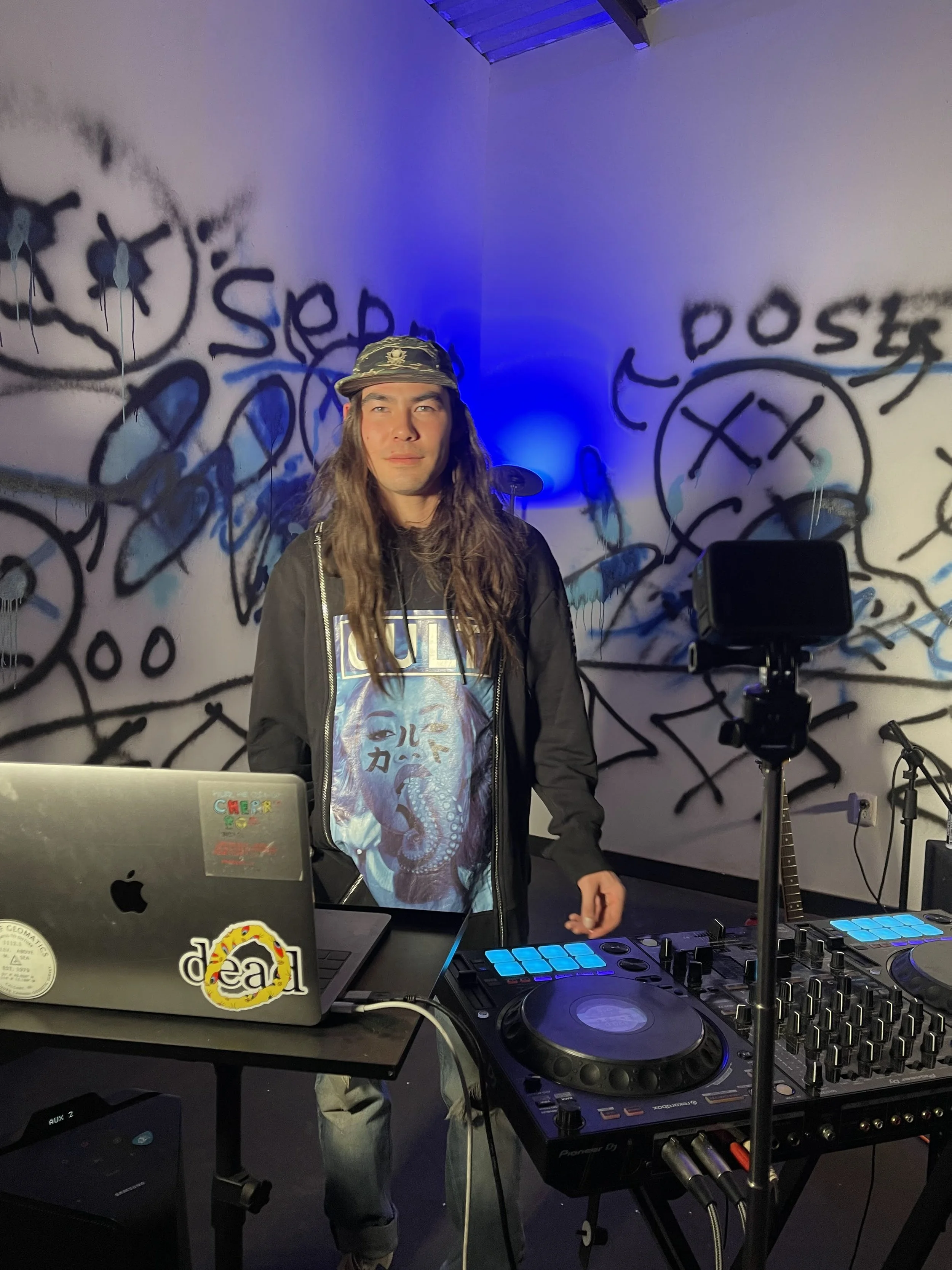 A young man with long hair wearing a baseball cap, DJ headphones, and a graphic T-shirt, standing behind DJ equipment with a laptop, controller, and microphone in a room with graffiti-style artwork on the walls and blue lighting.