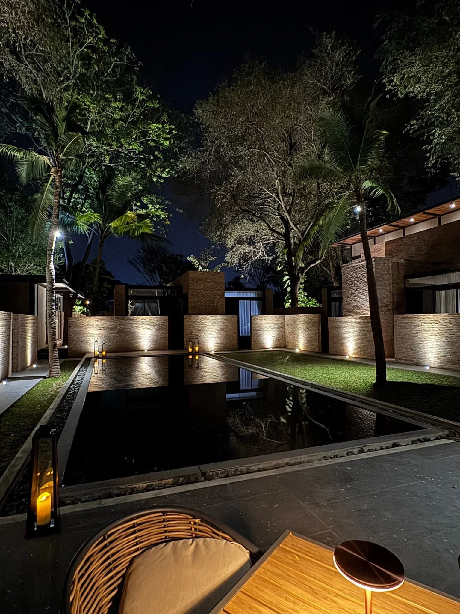 Makham Pool Access nighttime scene of an outdoor patio area with a rectangular swimming pool, tropical trees, and buildings with modern architecture illuminated with lights.