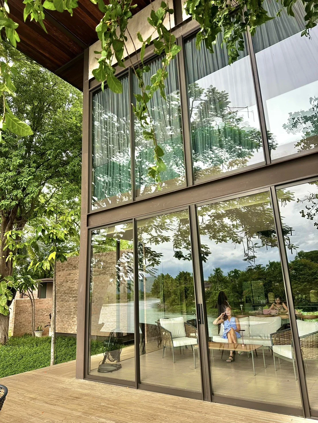 Modern house with large glass windows reflecting trees and sky, with people sitting inside and outdoor furnitures on a wooden deck overlooking by riverside overlooking River Kwai bridge, Kanchanaburi.