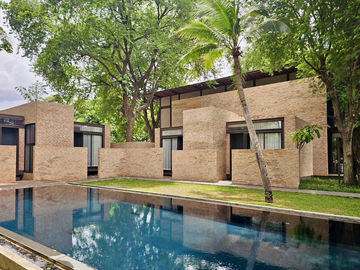 Modern brick residential building with large windows, surrounded by trees and greenery, with a swimming pool in the foreground and by riverside of River Kwai with a view overlooking River Kwai bridge.