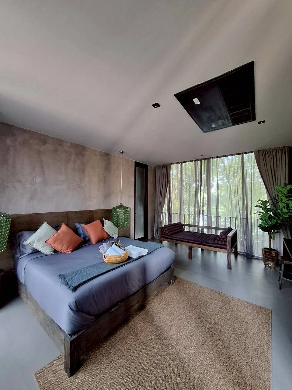 A Makham room with a king size bed, decorative pillows, a basket of toiletries, and a view of trees through large windows by River Kwai with curtains.