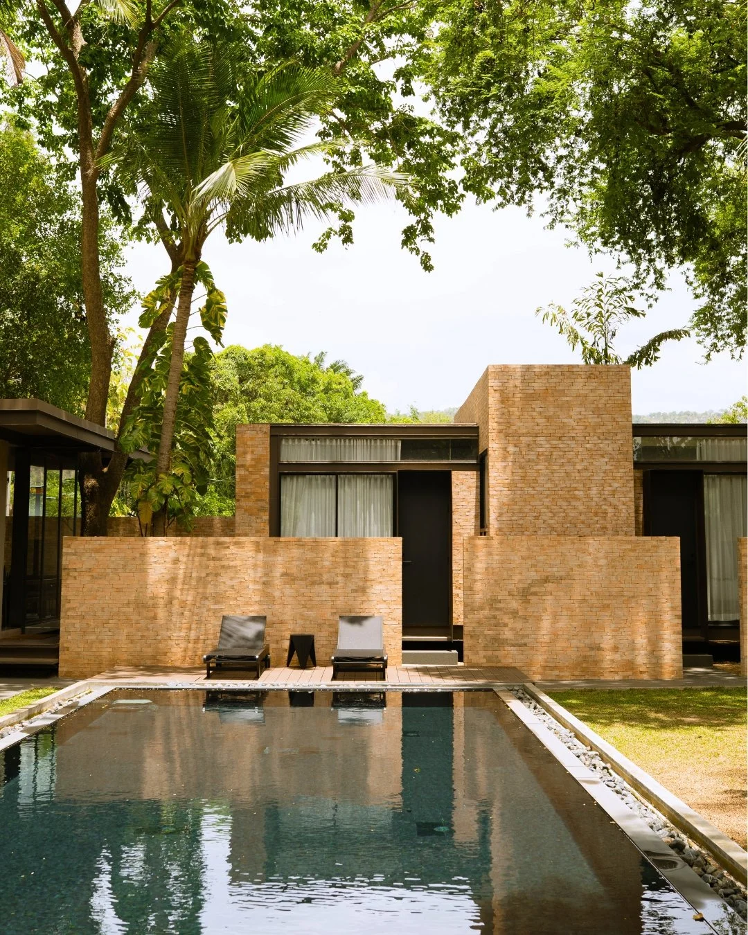 Modern house, Pool Access room, with brick facade and large glass windows, surrounded by lush green trees, with a swimming pool and swimming pool lounge chairs in the foreground.