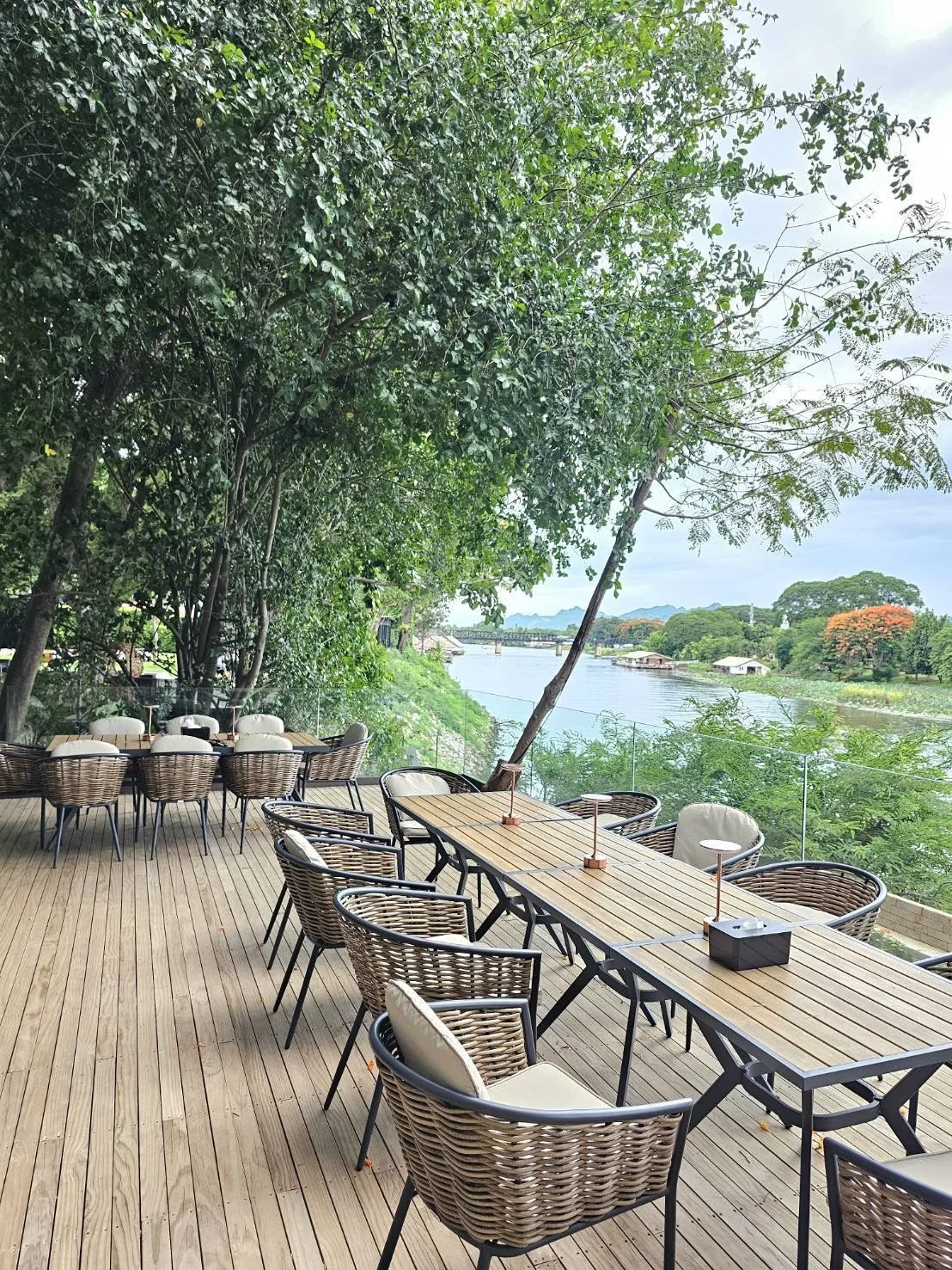 Outdoor balcony area with woven chairs and wooden tables by River Kwai and overlooking River Kwai bridge, surrounded by lush green trees.