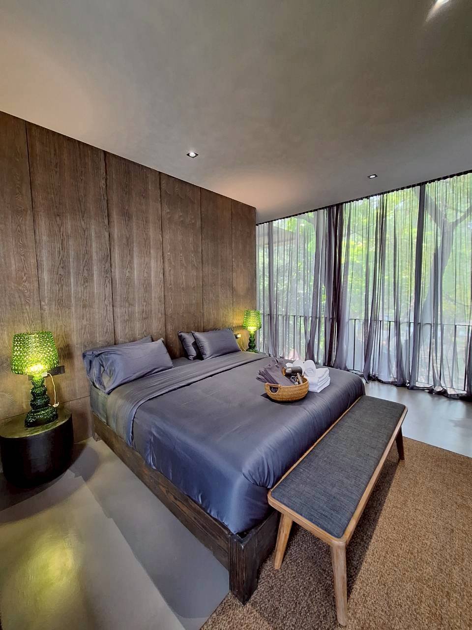 A modern Makham River View Suite bedroom featuring a wooden accent wall behind a gray bed with matching pillows and bedding. There are two green table lamps on black side tables on either side of the bed. A wooden bench with gray upholstery is positi